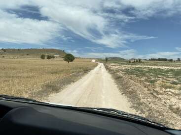 A dirt road stretches into the distance through dry fields under a bright blue sky with wispy clouds. The photo is taken from a vehicle.