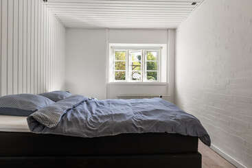 This is a minimalistic bedroom with white walls and ceiling, featuring a double bed with blue bedding, and a window letting in natural light.