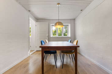 This image shows a minimalist dining room with a wooden table, four colorful chairs, wooden floor, white walls, a golden pendant lamp, and ample natural light.