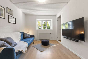 This cozy living room features a blue sofa, three pillows, wall-mounted TV, small window, framed pictures, pouf, rug, white walls, and wooden floors.