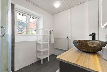 This bathroom features a stone vessel sink, wooden countertop, modern black faucet, white shelving, towel rack, large window, gray tile floor, and minimalistic white walls.