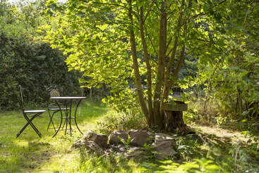 A peaceful garden scene with green foliage, sunlight streaming through trees, a metal table and two chairs, a stone circle, and a tree stump bench.