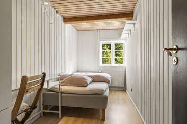 This is a small, cozy bedroom with wood-paneled walls and ceiling. There’s a single bed, a wooden chair, and a window letting in natural light.
