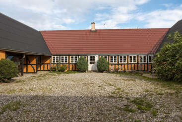 This image shows a traditional half-timbered farmhouse with a red-tiled roof, multiple windows, gravel courtyard, bushes, greenery, and outdoor furniture under a partly cloudy sky.