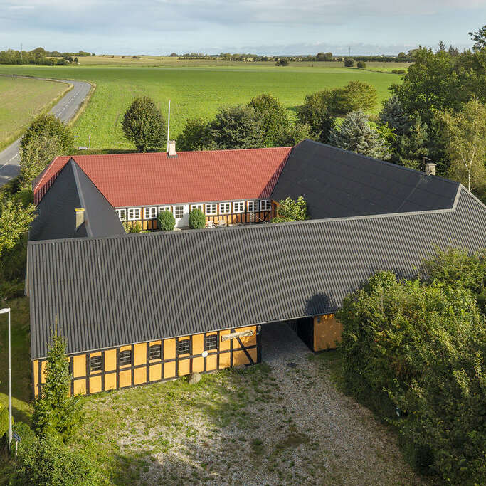 This image shows a large, L-shaped farmhouse with red and black roofs, surrounded by trees and fields, located beside a winding country road.
