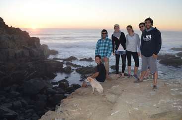 A imagem retrata um grupo de pessoas reunidas em uma praia, com alguns indivíduos se destacando, mostrando um ambiente descontraído e casual, provavelmente durante uma reunião ou evento social.