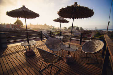 Um terraço de madeira na cobertura com cadeiras de vime, mesas redondas e três guarda-sóis de palha tem vista para uma cidade tranquila ao pôr do sol, criando uma atmosfera relaxante ao ar livre.