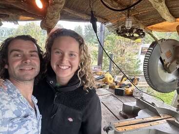 A man and woman smile together in a rustic outdoor workshop, surrounded by carpentry tools, wood, and greenery, creating a friendly and industrious atmosphere.