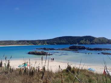 Eine ruhige Strandszene mit türkisfarbenem Wasser, Leuten, die schwimmen, sich sonnen und spazieren gehen, Felsvorsprüngen im flachen Meer, grünen Hügeln und einem klaren blauen Himmel.