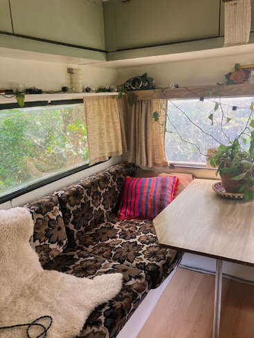 Esse interior aconchegante do trailer apresenta assentos com estampas florais, uma mesa de madeira, vasos de plantas, luzes de corda, janelas grandes com cortinas e uma almofada listrada colorida, criando uma atmosfera relaxante.
