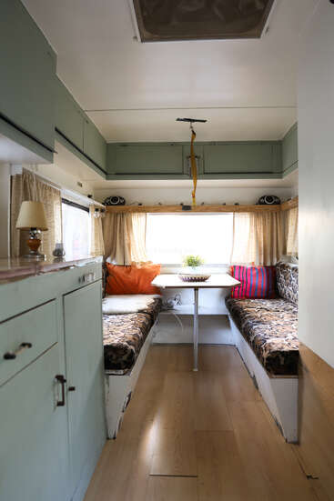 O interior desse campista aconchegante apresenta dois bancos estampados, uma pequena mesa, almofadas, armários de armazenamento superiores, piso de madeira, cortinas, uma lâmpada e uma planta decorativa.
