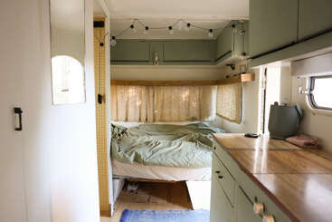 O interior desse aconchegante campista apresenta uma cama compacta com roupa de cama verde, cortinas amarelas macias, luzes de corda acima, armários verdes, bancadas de madeira e luz natural das janelas.