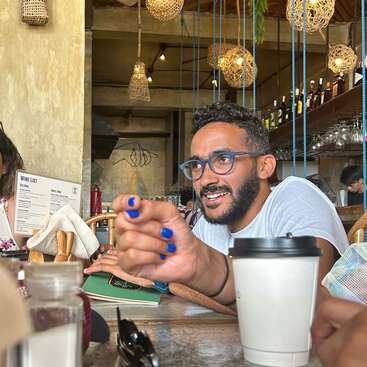 La imagen muestra a un hombre con las uñas pintadas de azul, sentado en la mesa de un restaurante, que gesticula hacia la cámara con la mano derecha. Lleva una camiseta blanca.