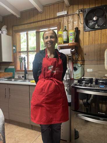 Eine Frau steht lächelnd in einer gemütlichen Küche und trägt eine rote Schürze. Holzwände, Fenster, Küchenutensilien, Herd und Flaschen verleihen dem Raum Wärme und Funktionalität.