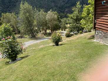 Ein üppiger, grüner Garten mit blühenden Sträuchern und Bäumen, einem gewundenen Kiesweg und einer Holzhütte inmitten einer malerischen, bewaldeten Berglandschaft im Sonnenlicht.
