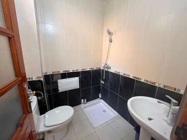 This image shows a compact bathroom with a toilet, sink, and shower area. The walls feature beige tiles above dark tiles, separated by a decorative border.