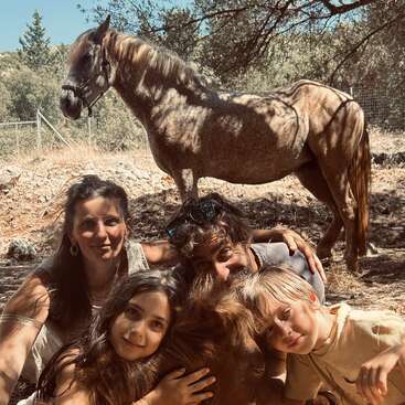 Four people, two adults and two children, smile and pose closely together outdoors under a tree with a brown horse standing behind them in the sun.