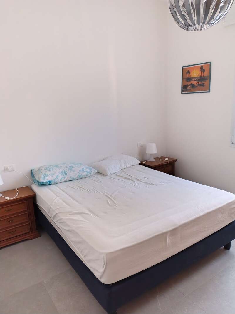 A simple, tidy bedroom with a double bed, two side tables, lamps, white walls, a ceiling light, and a framed sunset picture on the wall.