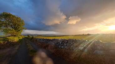 Uma cena rural pitoresca com um caminho estreito, um muro de pedra, campos verdes que se estendem ao longe, nuvens dramáticas e a luz dourada do sol a surgir por entre elas, criando uma atmosfera tranquila e serena.