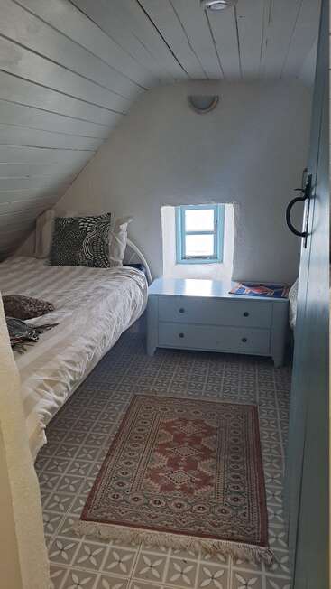 Um aconchegante quarto no sótão com uma cama de solteiro, almofadas decorativas, uma cômoda azul, uma janela pequena, um tapete estampado e um teto inclinado com painéis brancos. Um refúgio tranquilo.