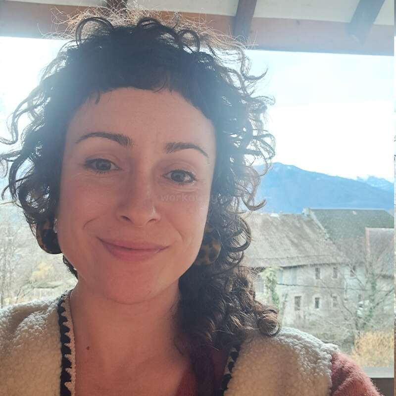Uma mulher de cabelos escuros e cacheados, usando brincos, sorri para a câmera. Ela está ao ar livre, com prédios rústicos e montanhas ao fundo, num dia ensolarado.
