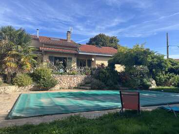 Detrás de un frondoso jardín hay una encantadora casa con tejado de tejas rojas. En el patio trasero, iluminado por el sol, hay una piscina cubierta y tumbonas. Ambiente tranquilo.