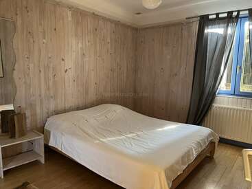 Esta imagen muestra un dormitorio sencillo con paredes revestidas de madera, una cama de matrimonio con ropa de cama blanca, una pequeña estantería, suelo de madera y una ventana con cortinas oscuras.