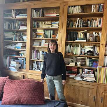 Una mujer frente a una gran estantería de madera llena de libros y cerámica, en el interior de una acogedora habitación con vigas de madera y cómodos asientos.
