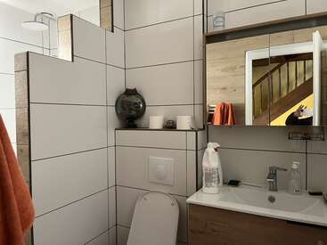 Esta imagen muestra un baño moderno con paredes de azulejos blancos, un inodoro, un lavabo, un armario con espejo, productos de limpieza y una toalla naranja que se refleja en el espejo.