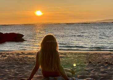 A person with long hair sits on sandy beach, gazing at the ocean during a golden sunset. Calm waves, rocks, and distant land complete the tranquil scene.