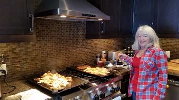 Uma mulher de cabelos longos e brancos, vestindo uma camisa xadrez vermelha, sorri enquanto prepara a comida no fogão de uma cozinha moderna com um revestimento de azulejos marrons.