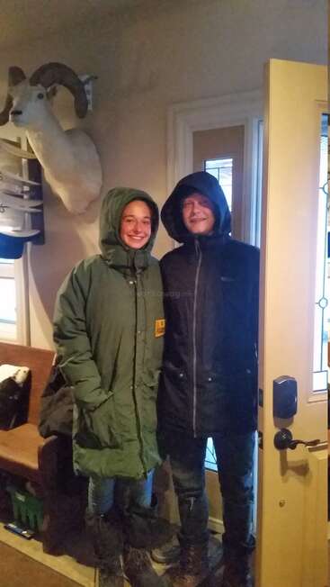 Duas pessoas vestindo casacos de inverno e botas estão em pé junto a uma porta aberta, sorrindo. Lá dentro, uma cabeça de carneiro empalhada está pendurada na parede atrás delas.