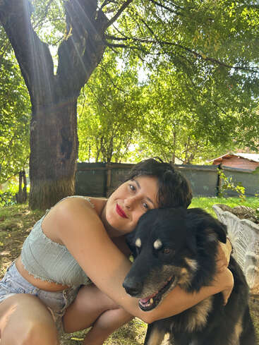 Uma jovem mulher de cabelos escuros curtos abraça um cachorro preto feliz ao ar livre. A luz do sol passa por entre as árvores, criando uma atmosfera calorosa, pacífica e afetuosa de verão.
