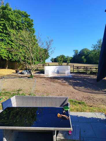 A sunny backyard scene with a glass table holding toys, wicker seating, bare dirt yard, leafy trees, white structure, and fields under a clear blue sky.