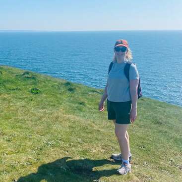 Una mujer está de pie en un acantilado cubierto de hierba junto al mar, con gafas de sol, gorra, camiseta gris, pantalones cortos y zapatos de montaña, disfrutando del tiempo soleado.