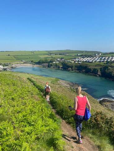 Two women walk along a narrow grassy path beside a turquoise river, with green fields, scattered houses, and clear blue skies in the background.