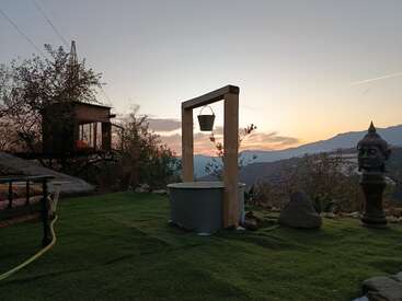 A serene garden scene at sunset, featuring a well with a bucket, a Buddha statue, a treehouse, green grass, and distant mountains under a colorful sky.