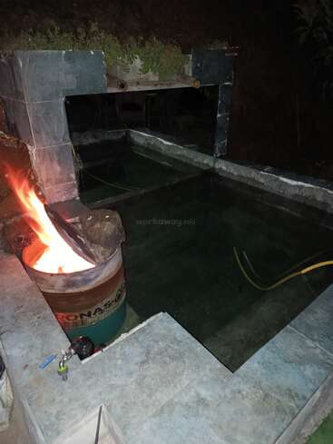The image shows a concrete outdoor hot tub or pool at night, with a barrel stove heating water, and pipes for circulation. There's greenery above.
