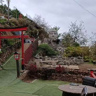 A peaceful outdoor setting features a large Buddha head statue on a rocky mound, Japanese torii gate, red lanterns, green artificial grass, and barrel table with chairs.