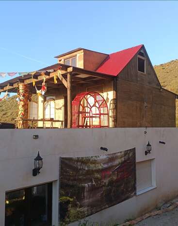 Une maison rustique en bois à deux étages, avec un toit rouge, des drapeaux colorés et un pilier décoré, trône au sommet d'un bâtiment blanc orné d'une fresque murale sur le thème de la nature.