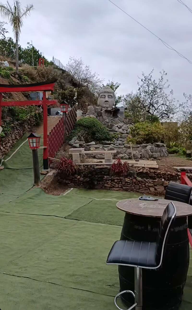 Un cadre extérieur paisible présente une grande statue de Bouddha sur un monticule rocheux, une porte torii japonaise, des lanternes rouges, du gazon artificiel vert et une table en tonneau avec des chaises.