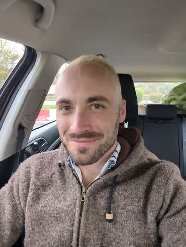 A man with short blond hair and a beard smiles while sitting in a car, wearing a brown zip-up hoodie. The background shows the car interior.