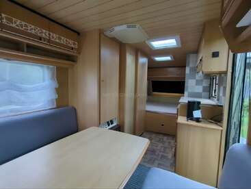 Esta imagen muestra el interior de una acogedora autocaravana, con muebles de madera, una mesa de comedor, asientos acolchados, una pequeña cocina, una zona de descanso y luz natural.