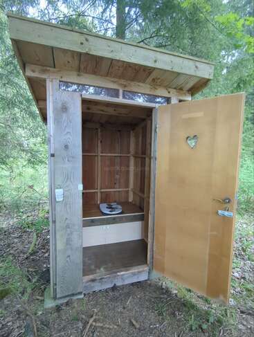 Esta imagen muestra una sencilla letrina de madera con la puerta abierta, en la que se aprecia un corazón recortado. Se encuentra en una zona boscosa, rodeada de vegetación.