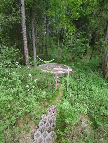 Una tranquila escena forestal con un sendero de piedra que conduce a una plataforma de madera. Hay una hamaca colgada entre los árboles, rodeada de una exuberante vegetación y flores silvestres.