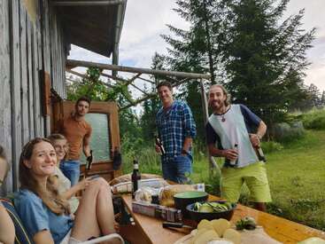 L'image représente un groupe de cinq personnes réunies autour d'une table en bois, dans un cadre extérieur serein avec des arbres et de l'herbe.