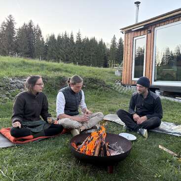 Three people sit on mats around a fire pit outdoors, talking and enjoying drinks. Behind them is a wooden cabin, surrounded by trees and grassy landscape.