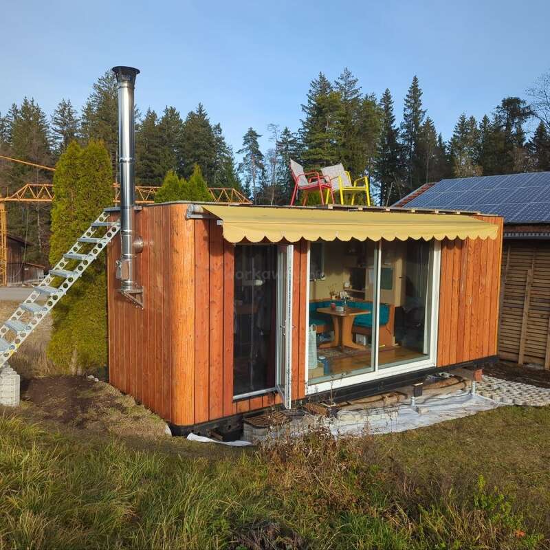 A small wooden tiny house with large glass doors, a metal chimney, and an awning. Colorful chairs sit on the roof; surrounded by trees and grass.