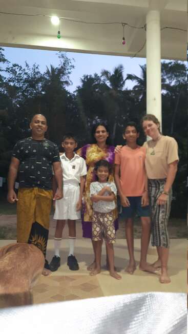 A group of six people stand together on a porch at dusk, smiling, with trees in the background and warm, casual clothing. A dog is in front.