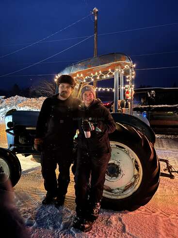 Two people dressed warmly stand in front of a decorated tractor at night, surrounded by snow. String lights illuminate the cozy winter scene, creating a festive atmosphere.
